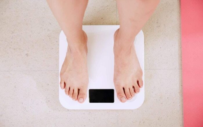 woman stepping on scale, woman looking at her phone and smiling, exercise equipment and fruit pictured against a wooden background, alarm clock, man enthusiastically lifting light weights, bowl of raspberries, personal trainer assisting a client in an exercise
